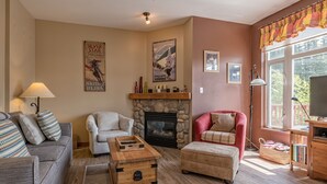 A cozy living room with a fireplace, a stone fireplace surround, a red armchair, a grey sofa, and a wooden coffee table. - Bright, upgraded townhouse in Creekside w/laundry, hot tub, close to village (Silver Star Mountain)