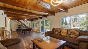 A spacious living room with a wooden ceiling, a glass coffee table, a brown sofa with decorative pillows, and a pool table. - Bare Necessities - AC, Hot Tub, Pool Table, and Lake Views! Walking distance to the Village. (Big Bear Lake)