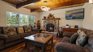 A cozy living room with a stone fireplace, leather sofas, and a large window offering a view of the outdoors. - Bare Necessities - AC, Hot Tub, Pool Table, and Lake Views! Walking distance to the Village. (Big Bear Lake)
