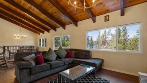A spacious living room with a large sectional sofa, a glass coffee table, and a chandelier. There are large windows providing a view of trees and a deck. - Bare Necessities - AC, Hot Tub, Pool Table, and Lake Views! Walking distance to the Village. (Big Bear Lake)