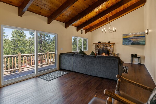 Un salón amplio con un sofá seccional gris grande, piso de madera, chimenea de piedra y un balcón con baranda de madera.
