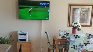 Un salon avec un téléviseur diffusant un match de golf, un coin repas avec des chaises et une table en bois, et un tapis à motifs sur le sol.
