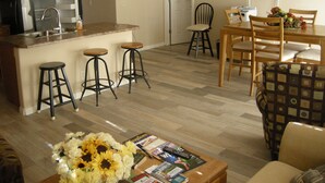 Une cuisine moderne dotée d’un réfrigérateur encastré, un coin repas avec des chaises et une table en bois, et un salon avec un canapé et une table basse.