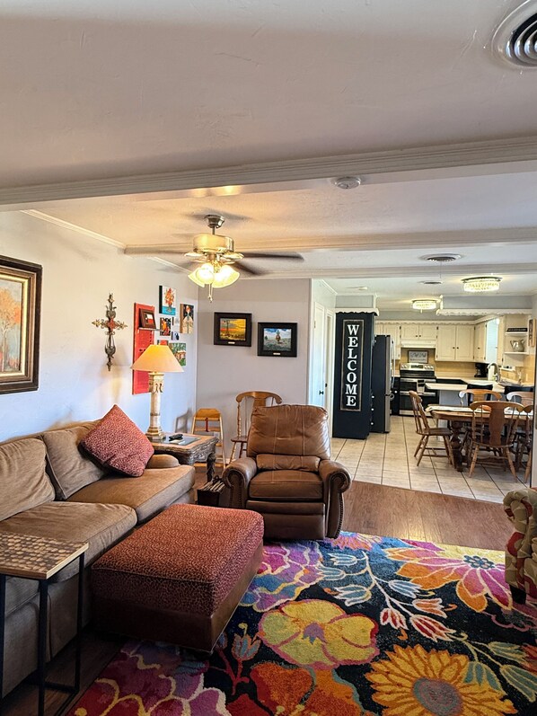 A living room with a sofa, ottoman, and a colorful rug. There is a dining area with a table and chairs, and a kitchen in the background.