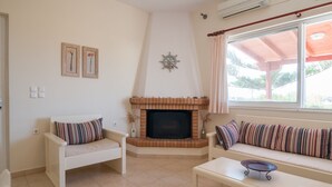 A living room with a fireplace, two white sofas with striped cushions, a small table, and a window with a view of greenery. - Felicia s sun-side house, Cycladic house by the beach, true owner hospitality (Naxos)