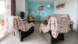 A modern living room with two sofas covered in patterned throw blankets, a small table, a chair, and a TV. The room has a teal accent wall and a window with curtains.