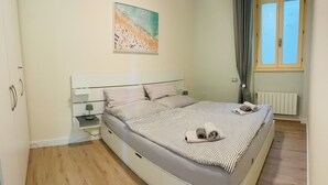 Une chambre à coucher comprenant un lit, une table de chevet, une lampe, un tableau, une fenêtre avec des rideaux et un radiateur.