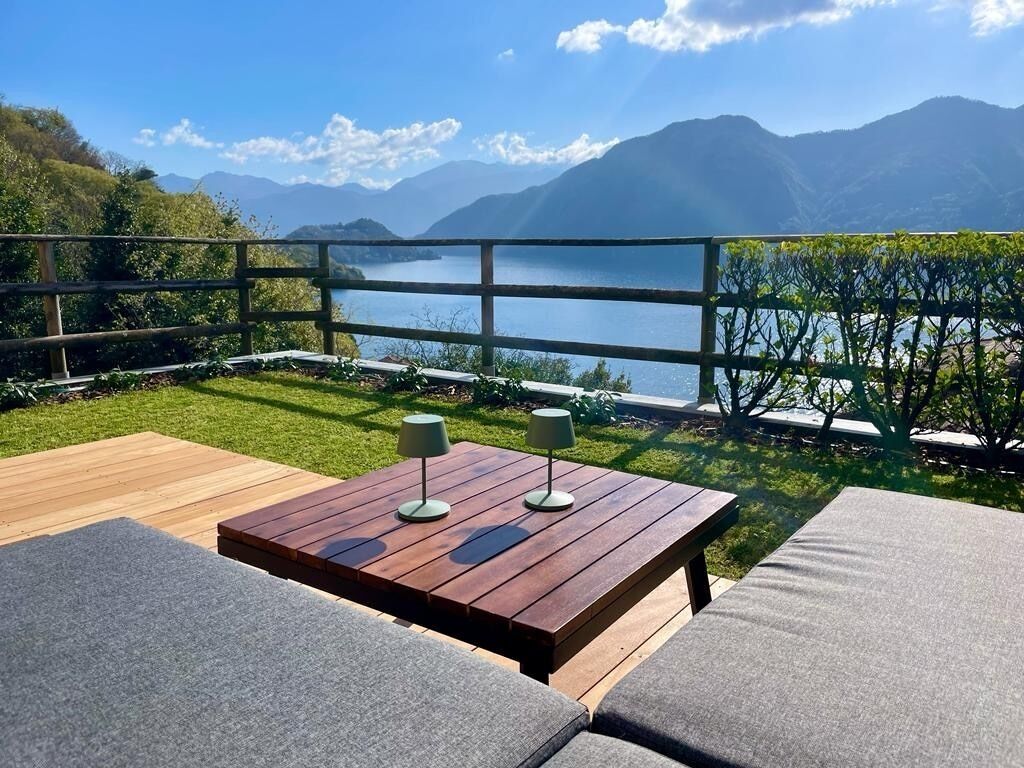 An outdoor seating area with a wooden table and cushioned chairs, overlooking a lake and mountains.