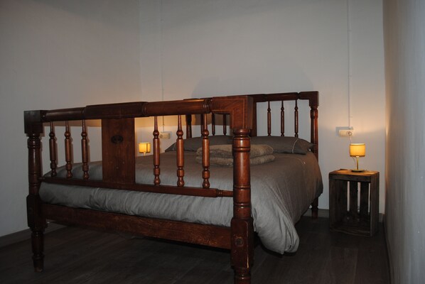 A wooden four-poster bed with a grey bedspread and pillows, a small bedside table with a lamp, and a dark wooden floor.