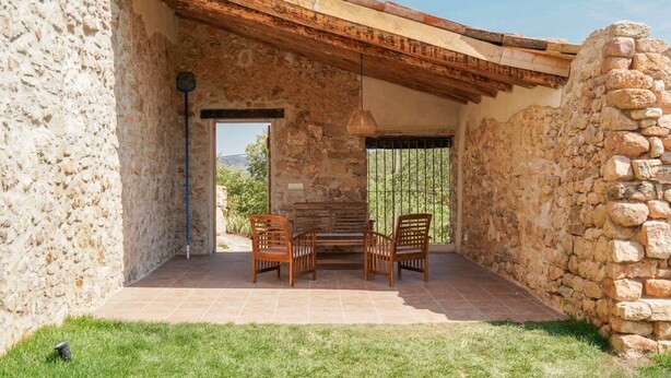 Un patio de paredes de piedra con dos sillas de madera y una mesa, un muro de piedra a un lado y vistas al exterior.