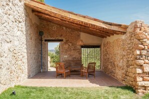 A stone-walled patio with two wooden chairs and a table, a stone wall on one side, and a view of the outdoors.