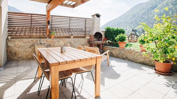 Un patio al aire libre con una mesa y sillas de madera, un muro de piedra, un horno de ladrillo y plantas en macetas.