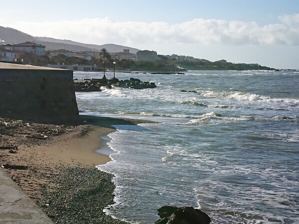 Un paysage côtier avec une plage de sable, des vagues qui s’écrasent sur les rochers et des bâtiments en arrière-plan.