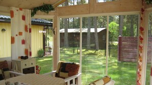 A wooden deck with a dining table and chairs, a glass door leading outside, and a view of a wooden cabin in a wooded area. - Lovely cottage in Juleboda, close to the sea with wonderful sandy beach (Degeberga)