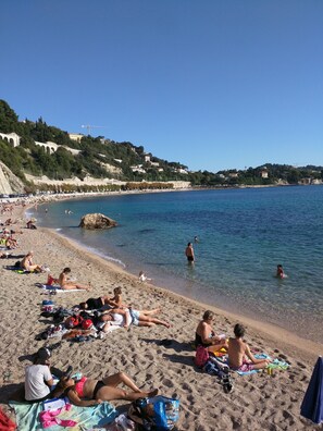 A beach with people sunbathing and swimming, a clear blue sky, and buildings in the background.