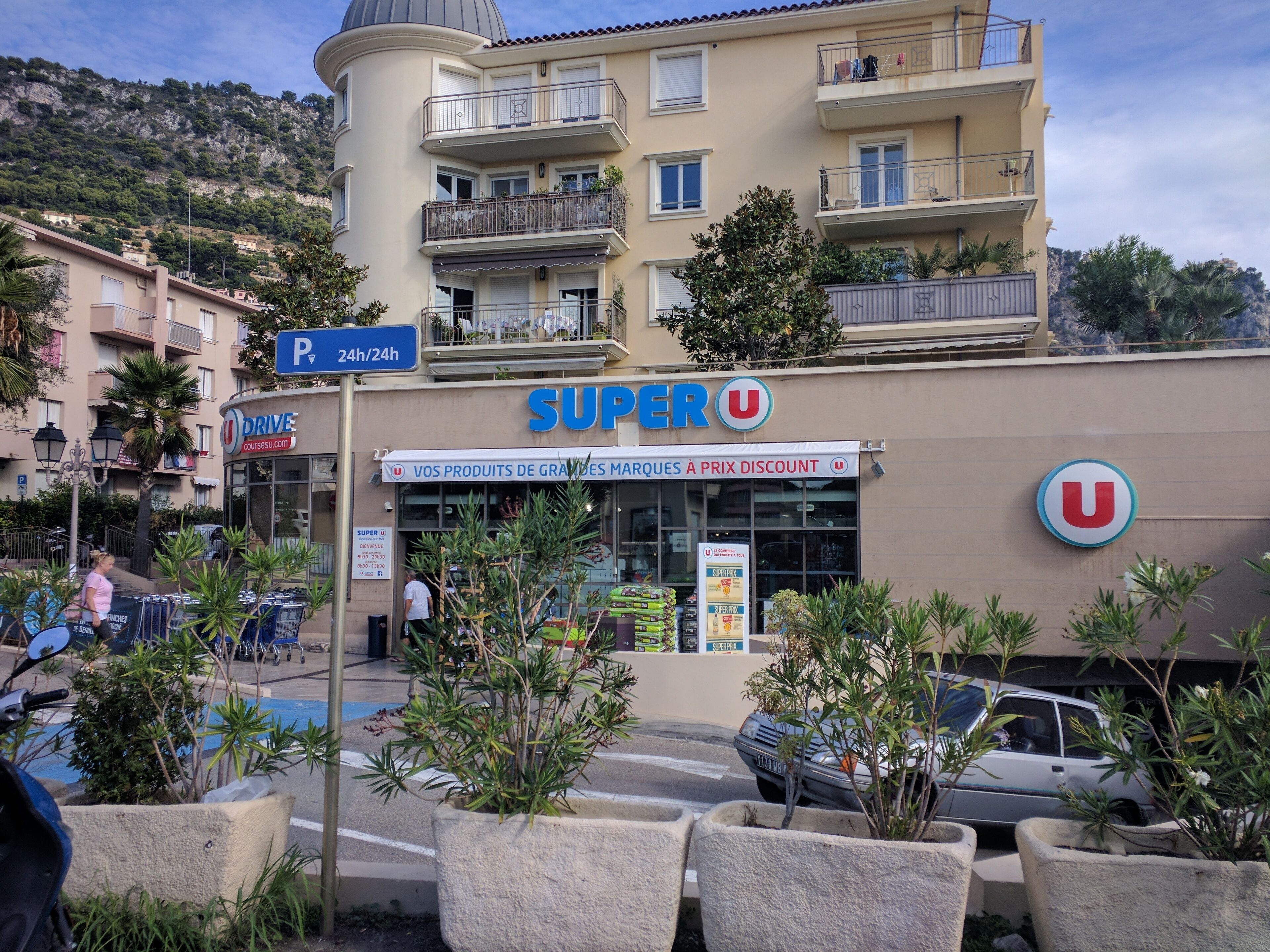 A Super U store with a parking sign, surrounded by potted plants and parked cars.