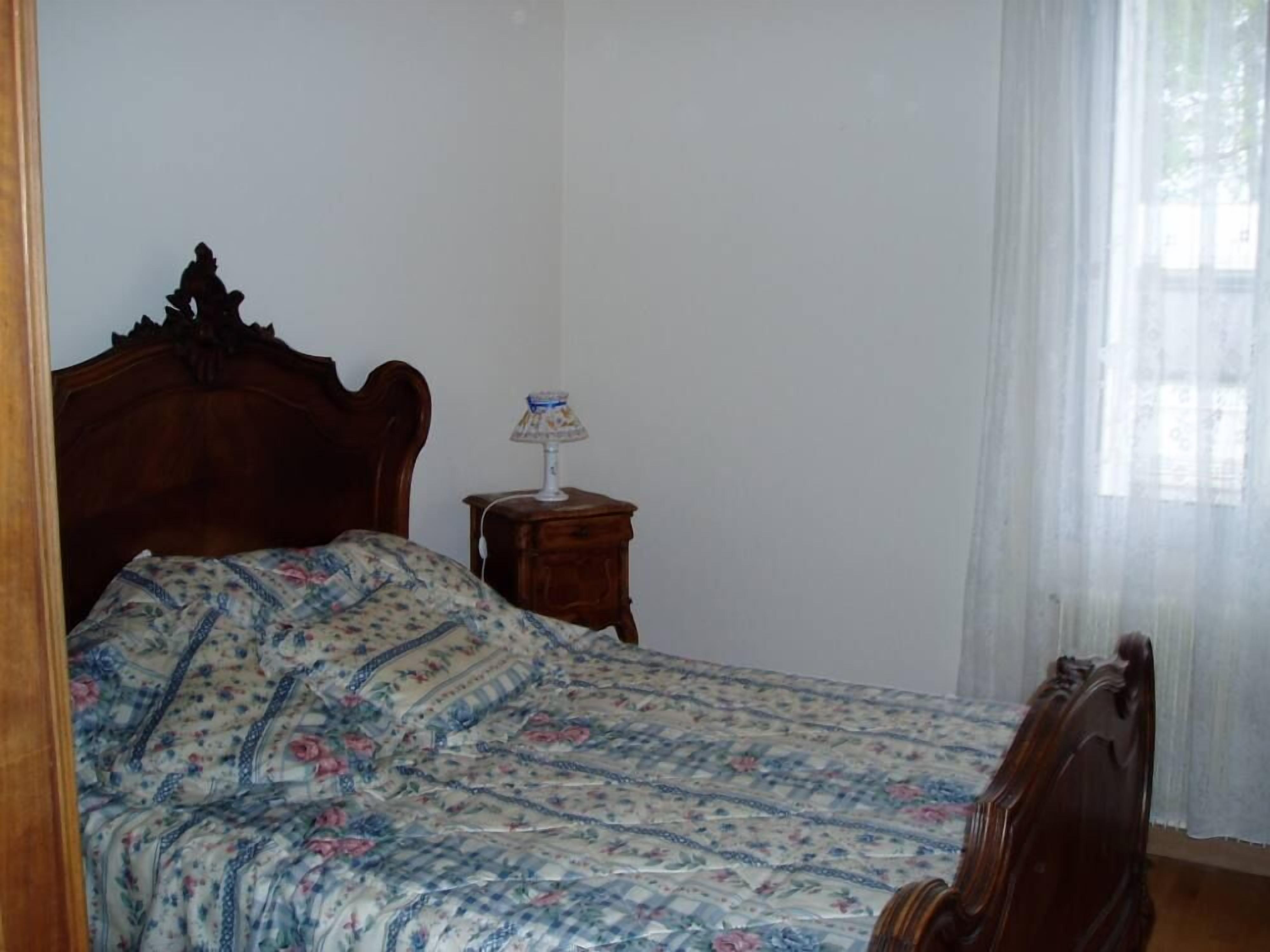 Ein Schlafzimmer mit einem hölzernen Bett, einem Nachttisch mit Lampe und einem Fenster mit transparenten Vorhängen.