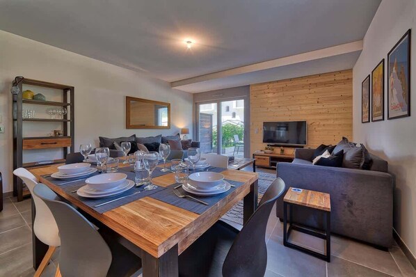 A modern living room with a dining area, a sofa, a TV, and a wooden wall. - Basecamp Town Centre 2 Bed Ground Floor Apartment with Terrace (Chamonix-Mont-Blanc)