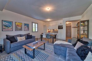 A modern living room with a sofa, a coffee table, and a dining area with chairs and a table. There are framed pictures on the wall and a kitchen area in the background. - Basecamp Town Centre 2 Bed Ground Floor Apartment with Terrace (Chamonix-Mont-Blanc)