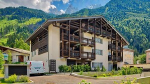 Un edificio de varios pisos con una estructura de balcón de madera, rodeado de abundante vegetación y montañas al fondo.