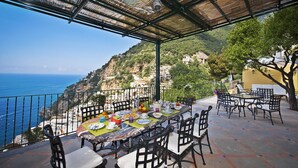 An outdoor dining area with a table set for a meal, overlooking a coastal landscape. - AMORE RENTALS - Villa Mimosa with Private Pool, Sea View, Air Conditioning, Positano Center (Positano)