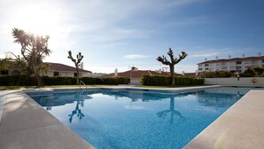 A swimming pool with clear blue water, surrounded by a concrete deck. There are palm trees and a building with a red roof in the background. - beautiful (Sitges)