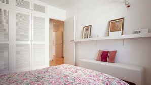 A bedroom with a floral bedspread, a white bench with a cushion, and a wall-mounted shelf with two framed pictures. - beautiful (Sitges)