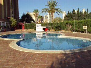 A swimming pool with a tiled deck, surrounded by palm trees and a building in the background. - Stunning 1 bedroom apartment with clear views across Benidorm (Benidorm, Alicante)