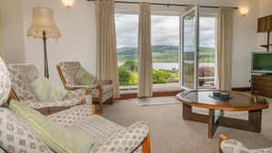 Una sala de estar acogedora con sofá, sillones, una mesa de centro y televisor. Amplias ventanas ofrecen una vista panorámica de vegetación y un lago.