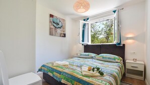 Une chambre à coucher comprenant un lit, une table de chevet, une chaise, une fenêtre avec des rideaux, un tableau mural et un luminaire suspendu.
