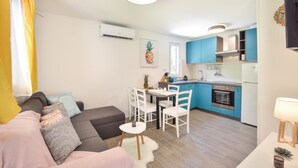 Un espace de vie compact comprenant un canapé, une table à manger, des chaises, un coin cuisine avec des meubles bleus et un petit réfrigérateur.
