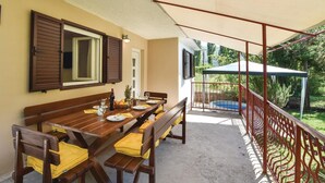 Un espace extérieur aménagé pour les repas, avec une table et des bancs en bois, un coin salon abrité et une vue sur une piscine et la verdure.