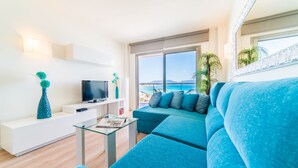 A modern living room with a large sofa, a glass coffee table, a flat-screen TV, and a view of the sea through large windows.