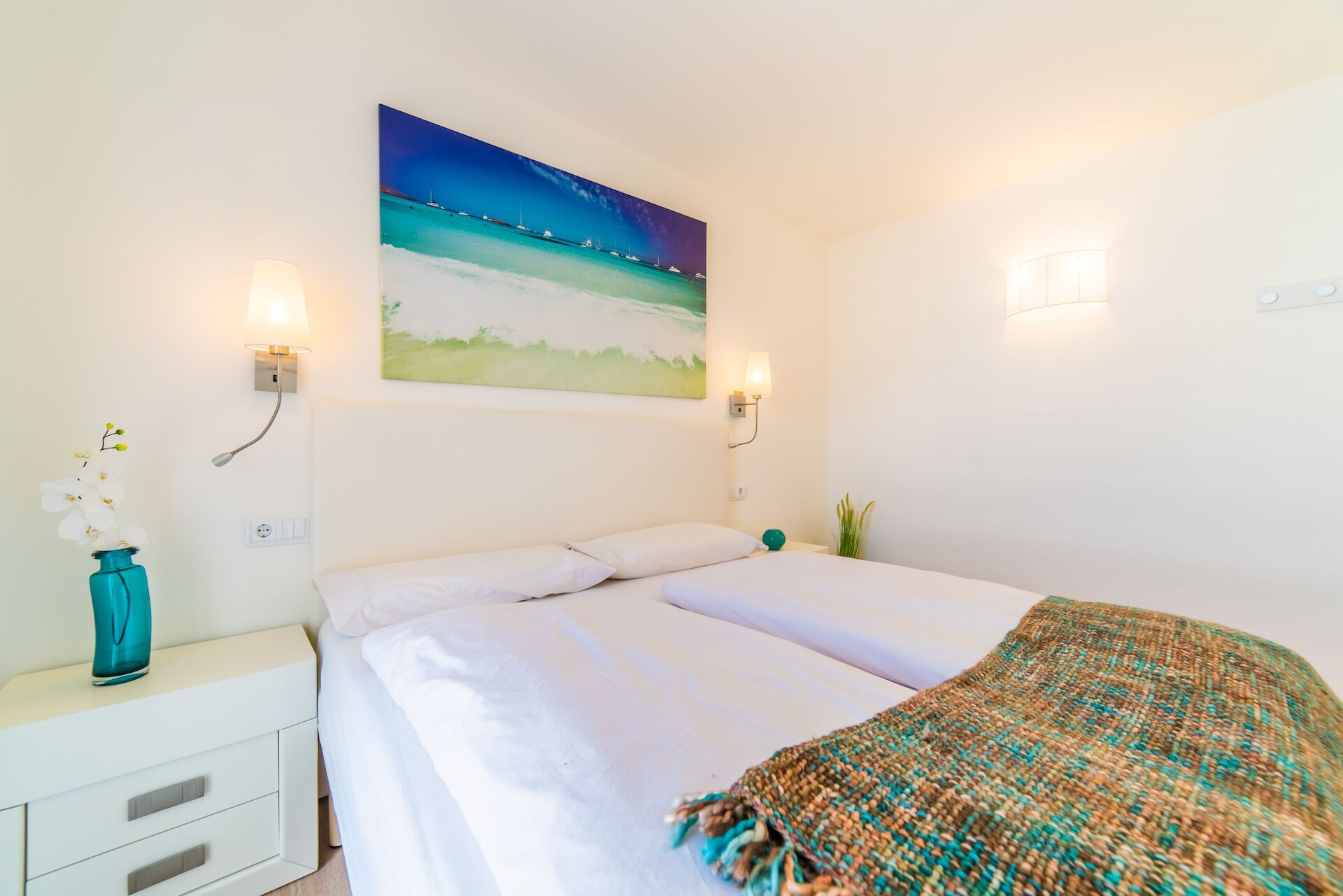 A hotel room with a bed, a nightstand, a vase with flowers, and a painting of a beach scene.