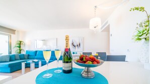 A modern living room with a white sofa, a blue accent wall, a dining area with a white table, a bottle of Moët & Chandon champagne, and a bowl of strawberries.