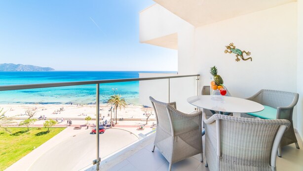 A balcony with a view of the beach and ocean, wicker chairs, a table, and a decorative frog wall art piece.