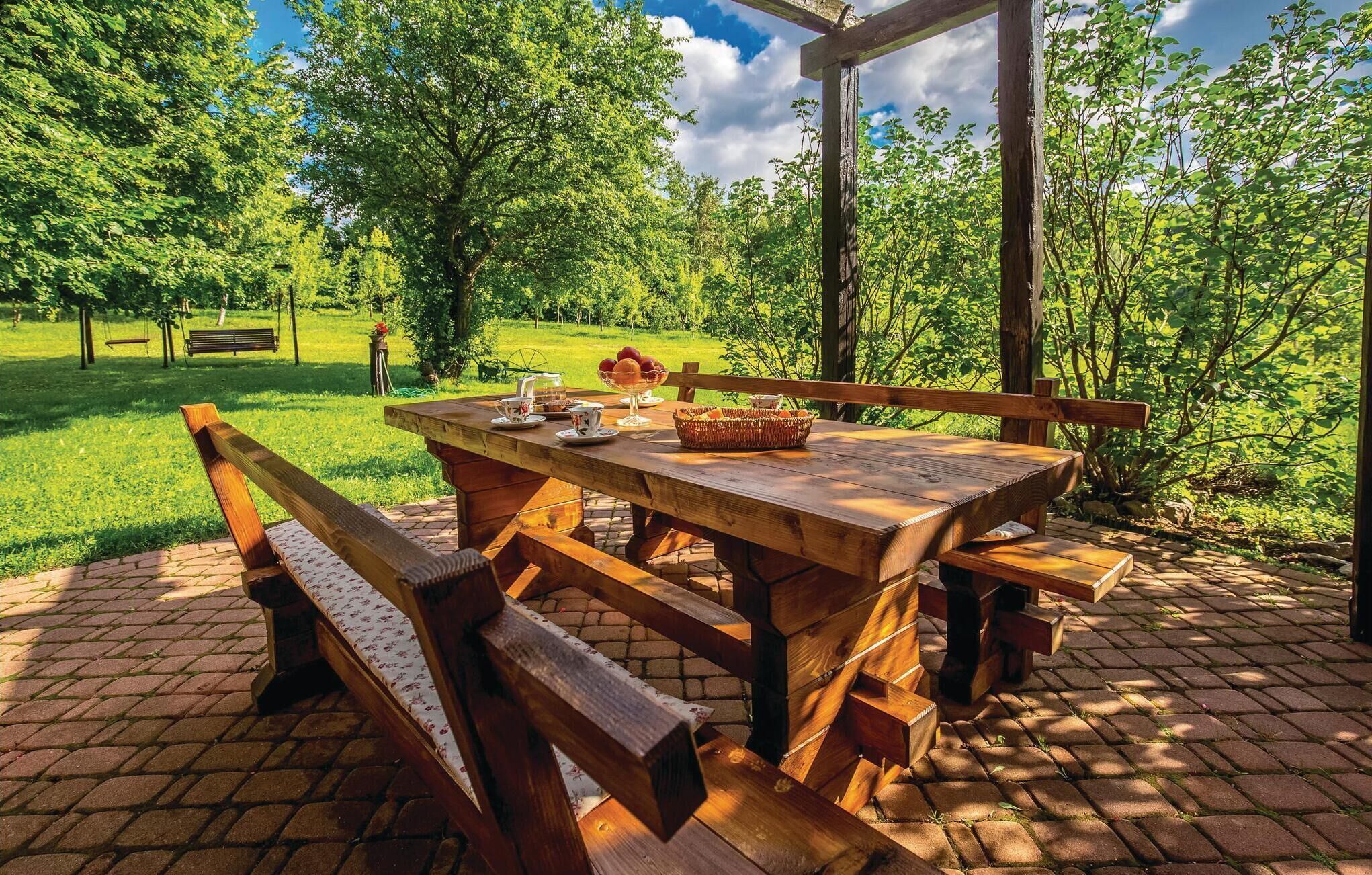 An outdoor wooden table with benches, surrounded by a brick patio, set in a lush green park with trees and a clear sky.