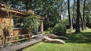 A wooden cabin with a glass door, a wooden deck, and a garden with trees and plants. - Gite with its private INDOOR swimming pool 30°  JACUZZI 36° private land 3000 m² (Naujac-Sur-Mer)