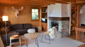 Een gezellige woonkamer met een stenen open haard, houten wanden, een bank en twee fauteuils.