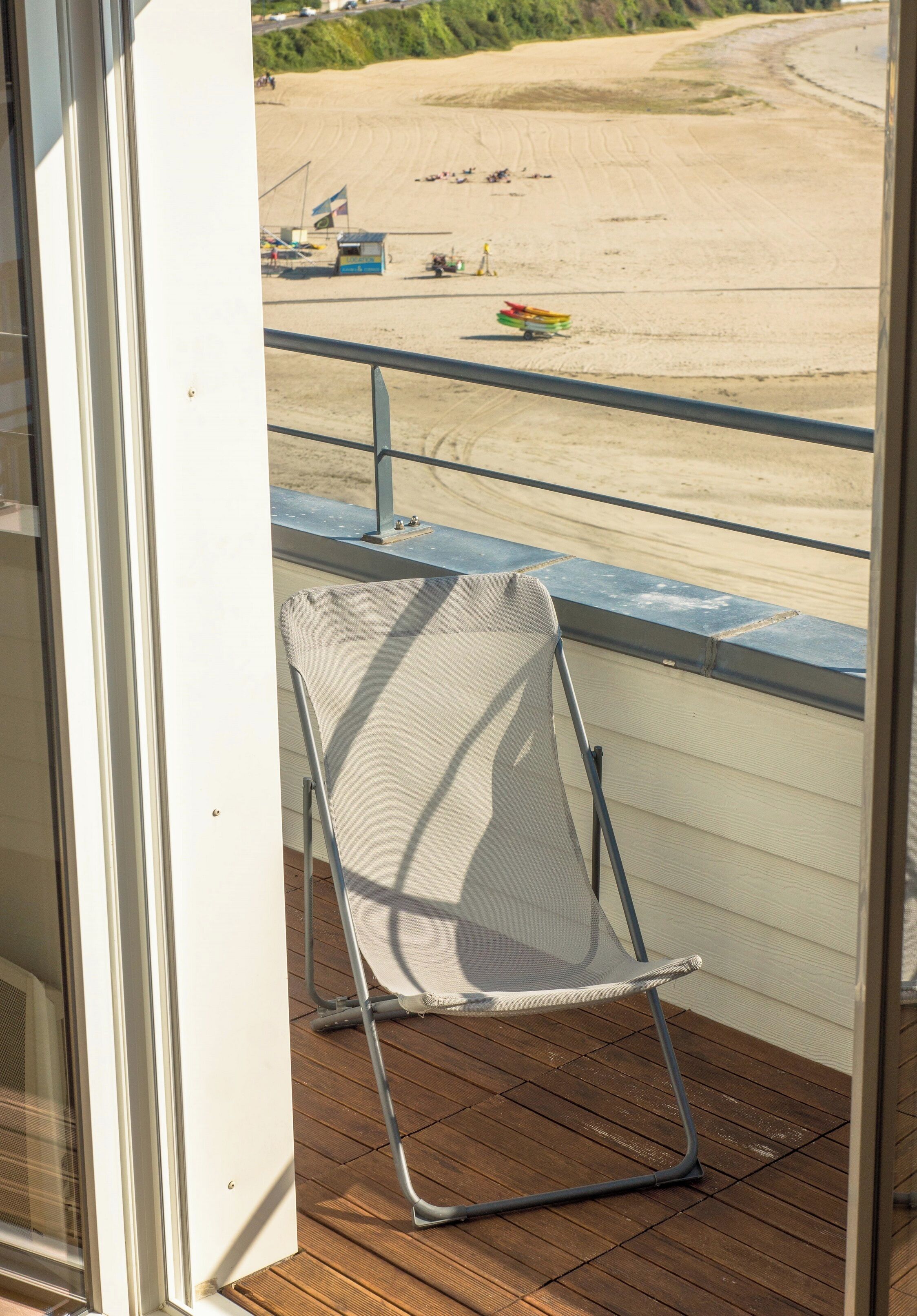Un balcón con una silla de metal y vistas a una playa arenosa con una bandera y una pequeña estructura.
