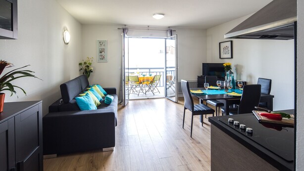 Un appartement moderne comprenant un espace de vie, un coin repas et une kitchenette. Il est équipé d’un canapé avec des coussins colorés, d’une table à manger avec des chaises et d’un téléviseur.