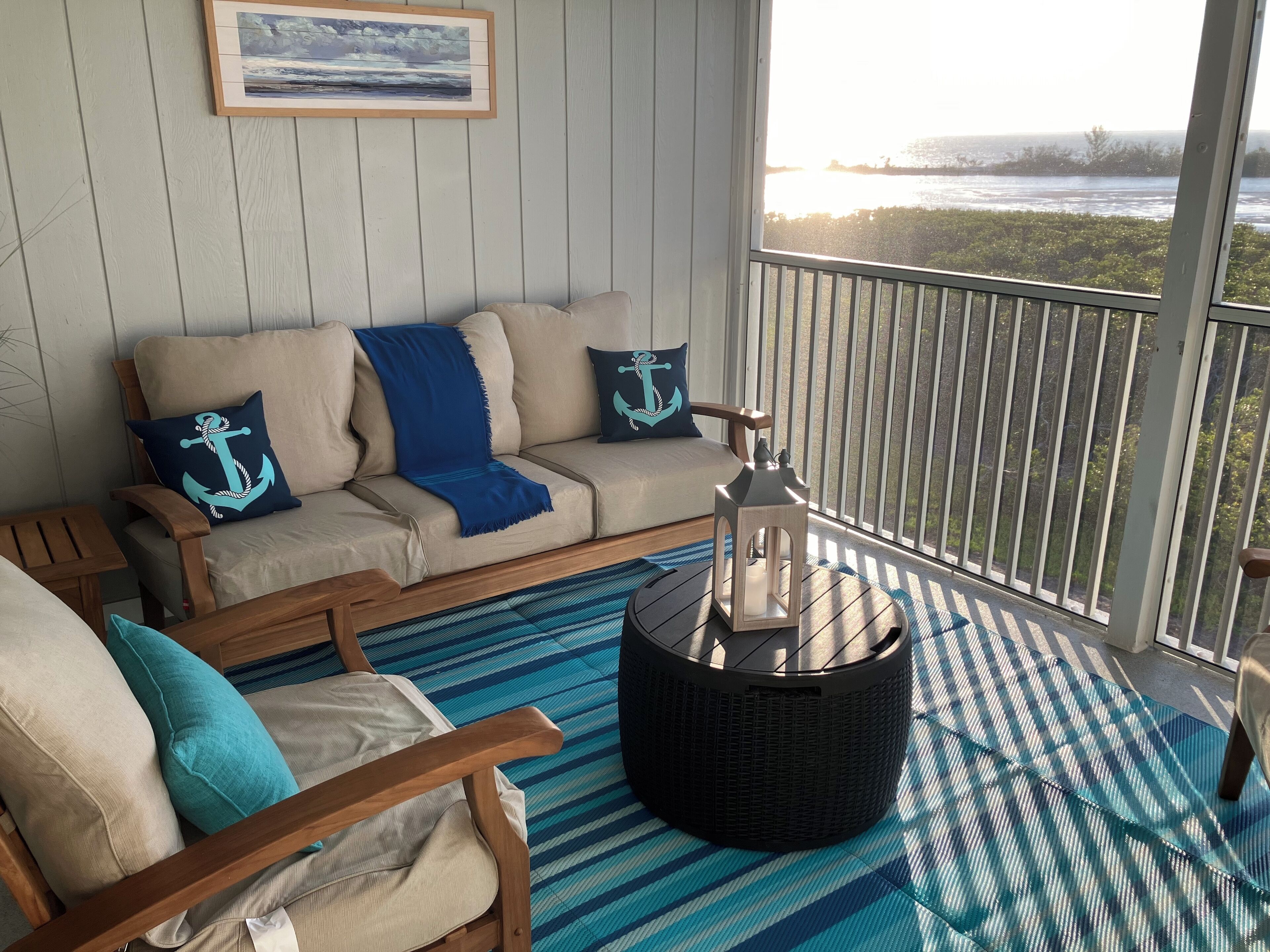 A cozy room with a beige sofa, two matching armchairs, a blue-striped rug, and a view of the ocean through a balcony railing.