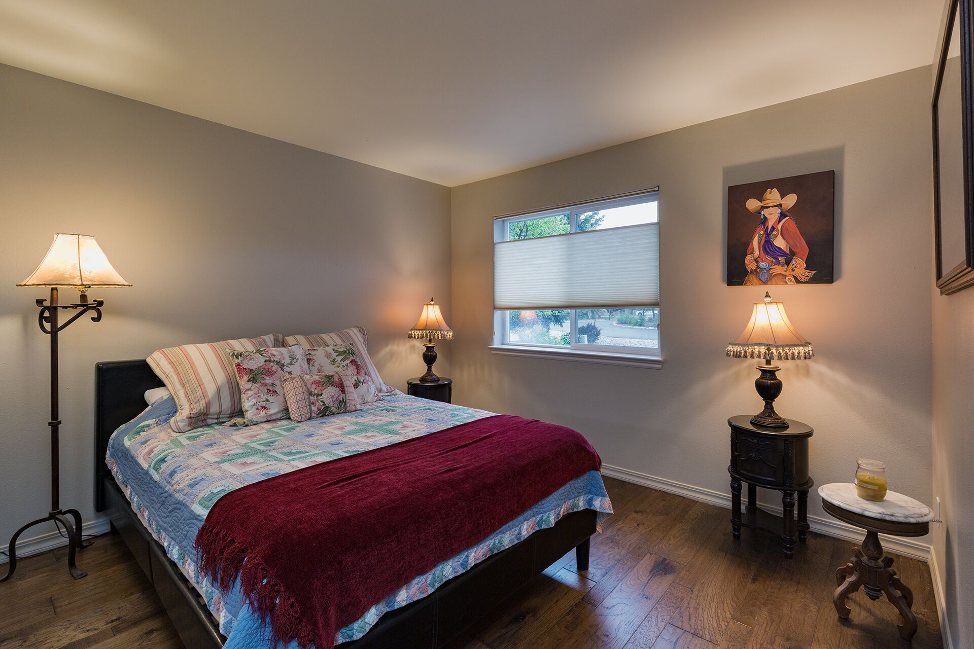 A bedroom with a bed, two bedside lamps, a window with blinds, a painting on the wall, and a small table with a lamp.