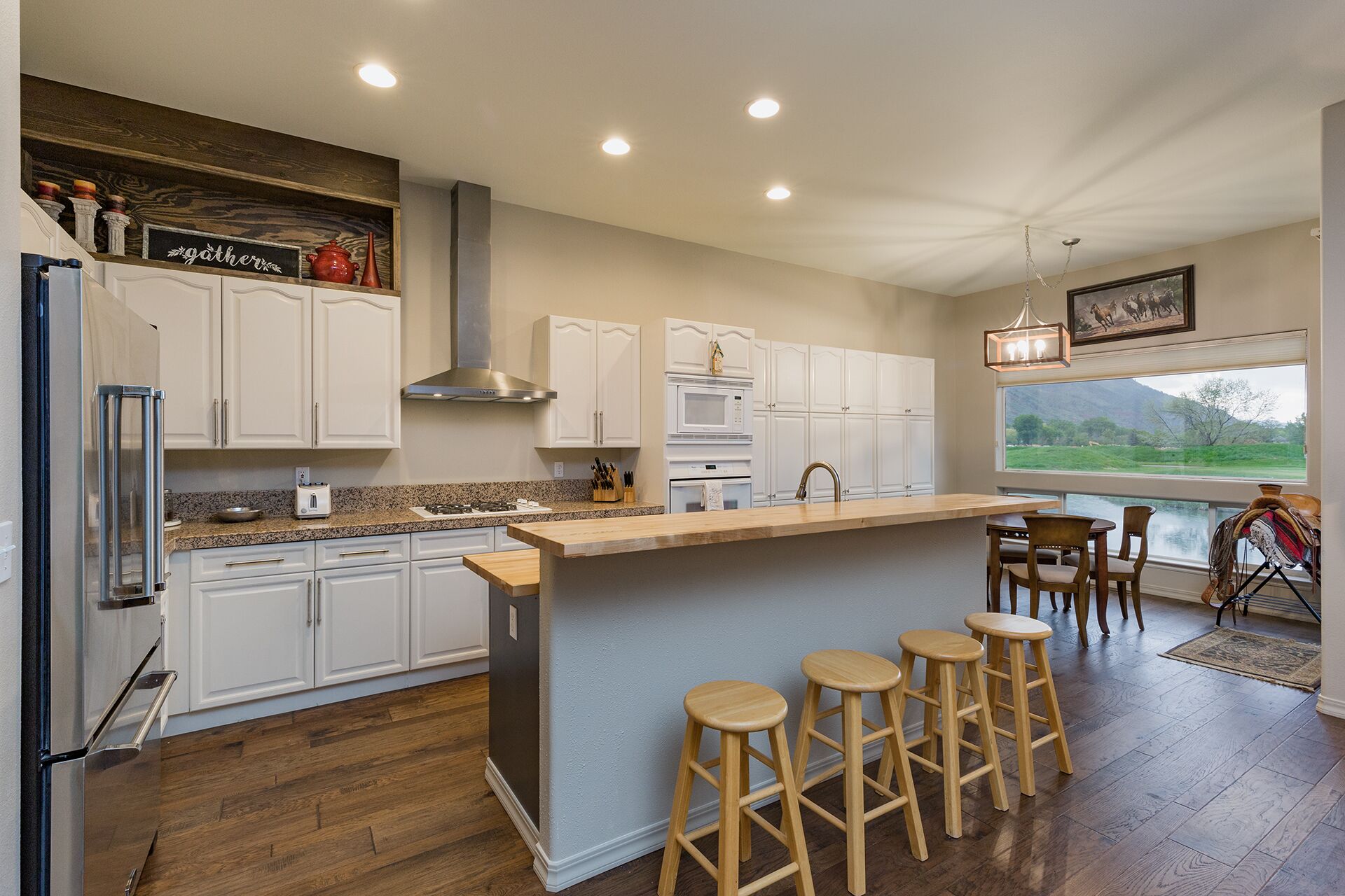 A modern kitchen with white cabinets, a central island with stools, a stainless steel refrigerator, and a view of a green landscape through a large window.
