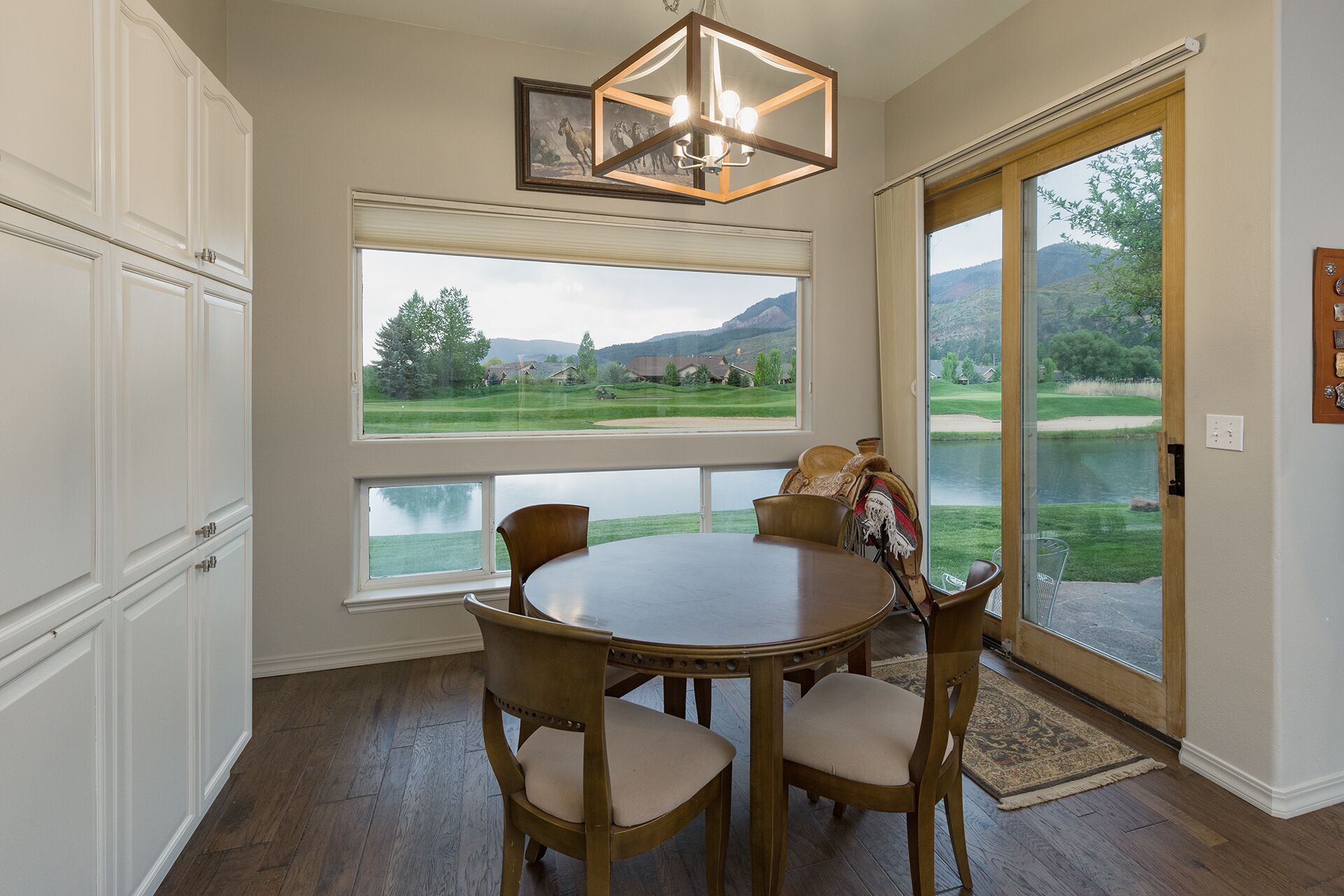 A dining area with a round table and four chairs, large windows with a view of a golf course and mountains, and a chandelier.