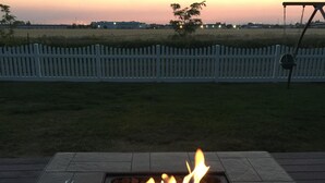 Ein quadratisches Feuerbecken mit Flammen, umgeben von Steinplatten, vor einem weißen Zaun und einem Sonnenuntergangshimmel.