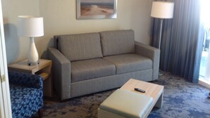 A hotel room with a grey sofa, a small armchair, a coffee table, a remote control, and a painting on the wall.