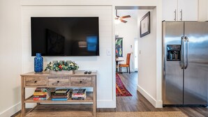Une cuisine moderne dotée d’un grand réfrigérateur en acier inoxydable, d’une console en bois sur laquelle sont posés des livres, d’un téléviseur à écran plat et d’un ventilateur de plafond.