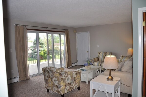 A living room with a sofa, armchair, coffee table, and lamp. There is a sliding glass door leading to a balcony. - Suttons Bay-across from Marina Park/Beach/By wineries, Brewery/Beautiful Sunrise (Suttons Bay)