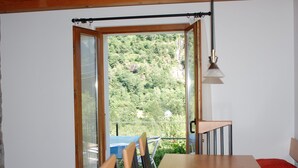 Une pièce avec une table et des chaises en bois, une fenêtre donnant sur une zone boisée et une lampe suspendue.